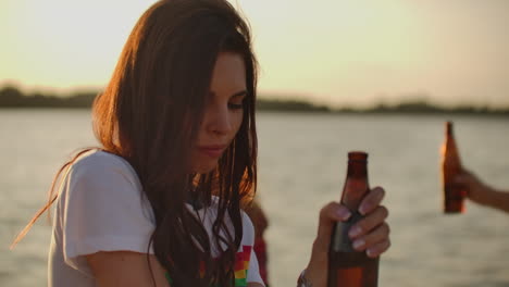 A-female-student-with-beautiful-gray-eyes-is-dancing-on-the-open-air-party-with-her-friends-and-beer.-She-smiles-and-touchs-her-long-dark-hair-and-enjoys-summertime-on-the-river-coast-at-sunset.
